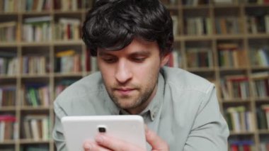 Evde tablet bilgisayar kullanan bir adam kitaplığın arka planında yatıyor.