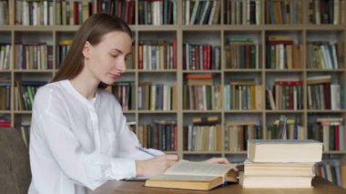 Üniversite kütüphanesinde kitap ve dizüstü bilgisayarla masada oturan ve okuyan genç bir kadın.