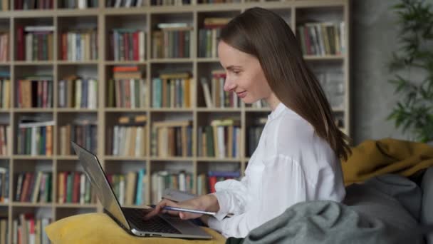 Mujer con documentos usando un portátil en casa sobre el fondo de un estante de libros — Vídeo de stock