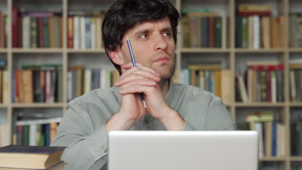 Zamyšlený mladý muž sedí u stolu v kanceláři, v ruce drží tužku, dívá se stranou, odpočívá od práce na notebooku, přemýšlí o plánu projektu — Stock video