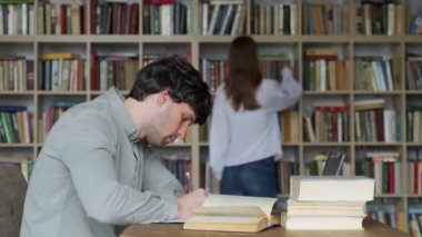 Erkek öğrenci kütüphanede sınav hazırlıyor ve dersler öğreniyor. Üniversite kütüphanesinde kitap ve dizüstü bilgisayarla masada oturan genç bir adam.