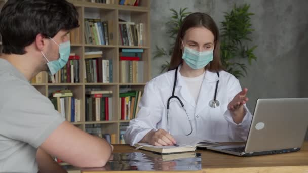 La doctora que usa una máscara médica se sienta en su escritorio y habla con un paciente mientras observa los resultados de sus pruebas mostrándolos en una computadora portátil. — Vídeos de Stock