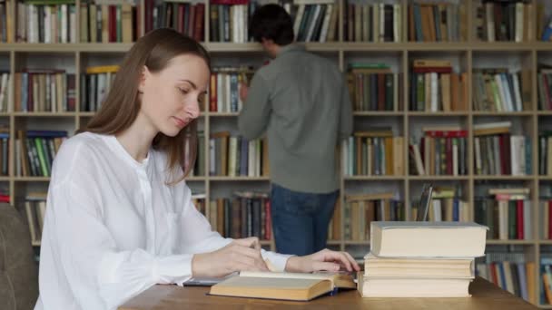 Jonge vrouw zit aan tafel met boeken en laptop in universiteitsbibliotheek en studeert — Stockvideo