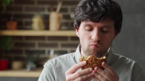 Hungriger Mann isst Burger in der heimischen Küche. Mann genießt Cheeseburger — Stockvideo