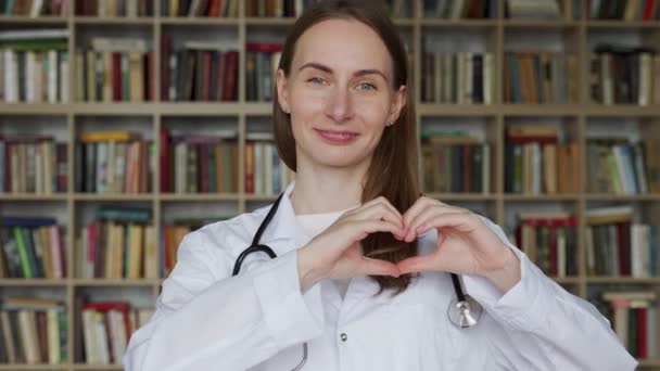 Giovane medico donna che fa segno di cuore con le mani sullo sfondo di una libreria — Video Stock