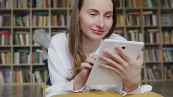 Młoda kobieta leżąca na podłodze za pomocą tabletu w domu na tle półki na książki — Wideo stockowe