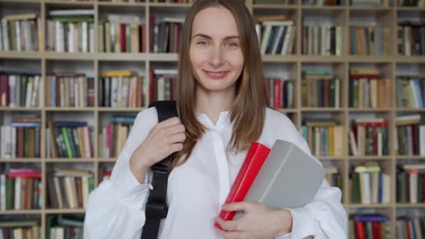 Młoda uśmiechnięta kobieta stoi z książkami i plecakiem w bibliotece — Wideo stockowe