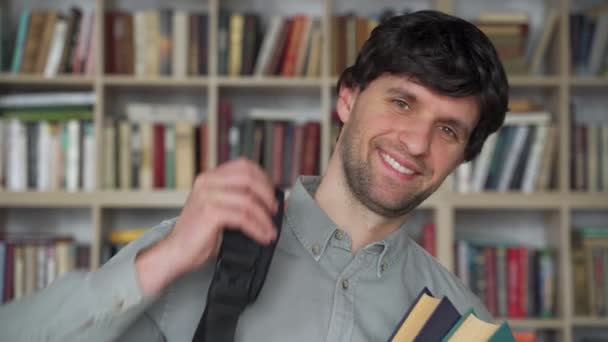 Junger Mann steht mit Büchern und Rucksack in der Bibliothek — Stockvideo