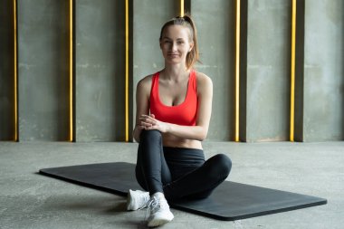 Sporcu kadın paspasın üzerinde oturuyor, kameraya bakıyor ve eğitim amacından bahsediyor.