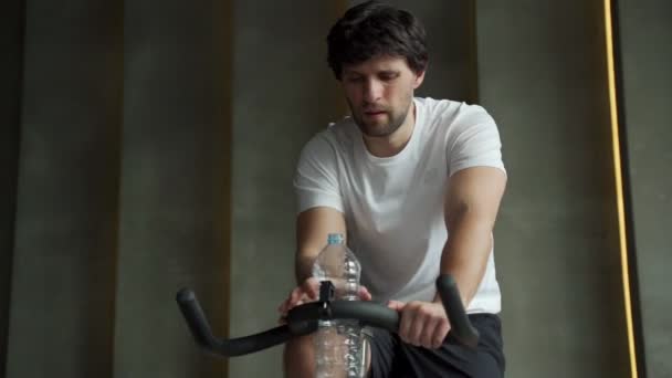 Unge man dricker vatten i gymmet. Idrottaren tränar på motionscykel på gymmet. — Stockvideo