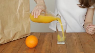 A woman pours orange juice into a glass and drinks it while sitting in the kitchen