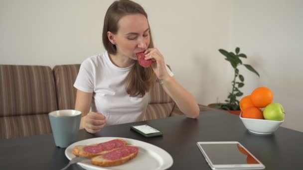 Frau mit Smartphone zu Hause in einer modernen Küche. Eine Frau isst ein leckeres Wurstsandwich — Stockvideo