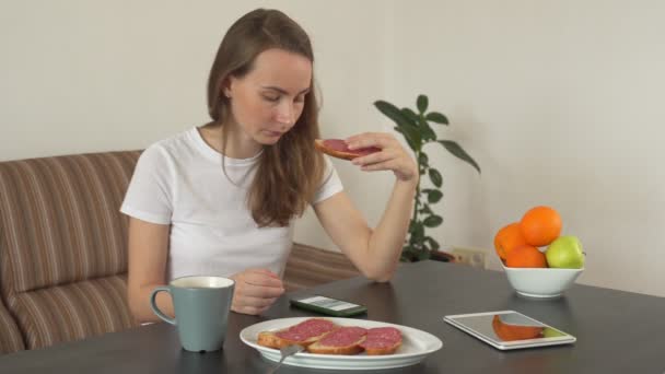 Frau mit Smartphone zu Hause in einer modernen Küche. Eine Frau isst ein leckeres Wurstsandwich — Stockvideo