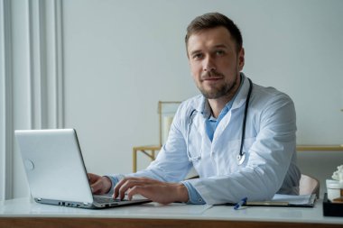 Tıp bürosunda dizüstü bilgisayar kullanan bir doktor. Adam tıbbi ofisinde belge ve dizüstü bilgisayarla çalışıyor.