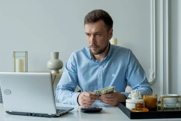 El hombre con dinero y una calculadora cheques facturas, calcula los gastos, estudia el saldo de crédito sentado en la mesa en casa — Foto de Stock