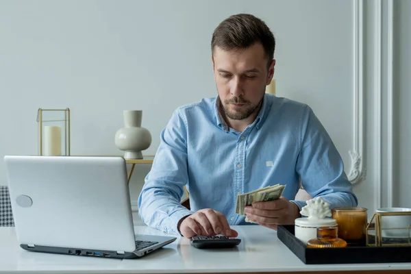 El hombre con dinero y una calculadora cheques facturas, calcula los gastos, estudia el saldo de crédito sentado en la mesa en casa — Foto de Stock
