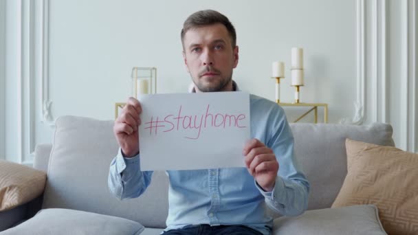 Человек, сидящий на диване предупреждает о коронавирусной болезни держит плакат остаться дома — стоковое видео
