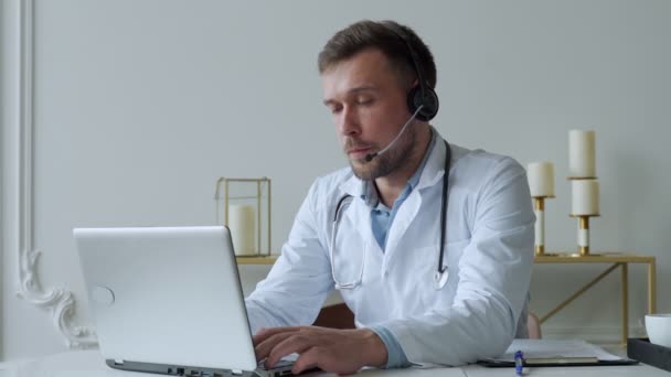 Retrato de hombre médico en bata blanca hablando en videollamada en auriculares en el ordenador portátil sentado en el gabinete que tiene consulta en línea con el paciente. — Vídeo de stock