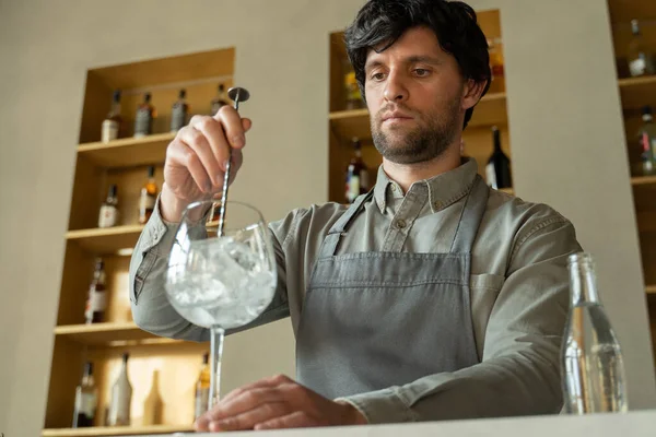 Önlüklü bir barmen barın arka planında kokteyl bardağındaki buzları karıştırıyor. — Stok fotoğraf