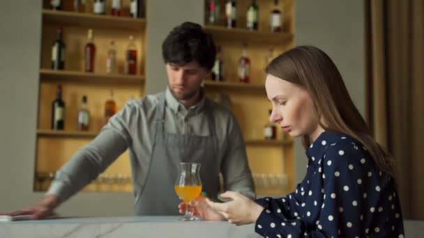 Frau mit Smartphone an der Bar vor dem Hintergrund eines Barkeepers, der den Tisch wischt — Stockvideo