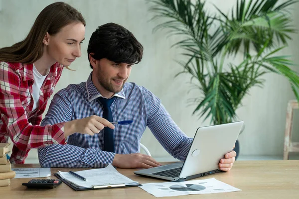 年轻夫妇在办公室一起工作。年轻女人笑着指着笔记本电脑，和同事谈论着什么 — 图库照片