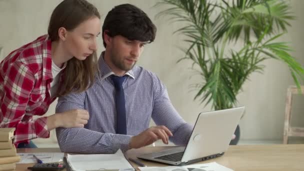 Молодая пара работает вместе в офисе. Молодая женщина указывает на ноутбук с улыбкой и обсуждает что-то со своим коллегой — стоковое видео