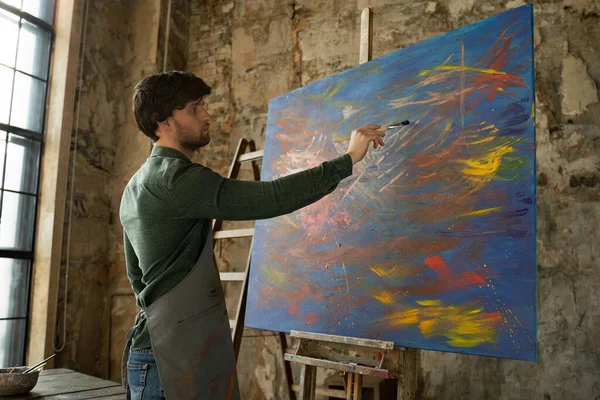 Mężczyzna artysta maluje obraz w studio. Portret mężczyzny malującego obraz niebieską farbą w warsztacie. — Zdjęcie stockowe