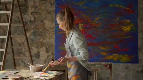 Resim stüdyosunda boya fırçasını büyük tuvalde tutan kadın sanatçı. Tuval üzerinde modern sanat eserleri — Stok video
