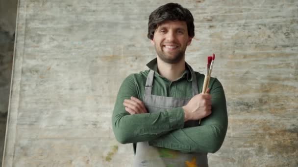 Portret van een tevreden jonge mannelijke kunstenaar die zijn armen kruist met een kwast in zijn hand op een achtergrond van zijn nieuwe schilderij — Stockvideo