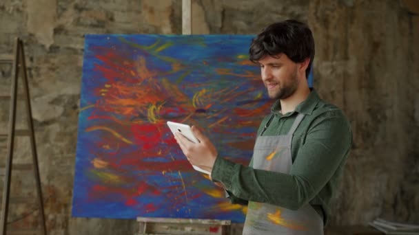 Erkek sanatçı stüdyosunda duruyor ve bir tablet kullanıyor, bir proje üzerinde çalışıyor. Yağlı boyalı atölye — Stok video