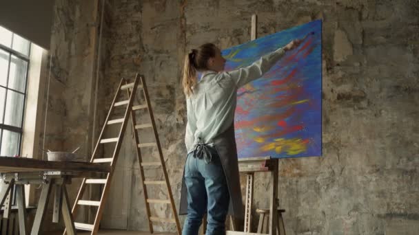 Portrait of young female artist wearing apron in art studio. Artist expressively paints a picture with a brush — Stock Video