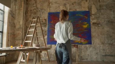Dişi ressam ellerini büyük tuvale koyup yağlı boya resim çizer. Ressam soyut sanat yaratıyor