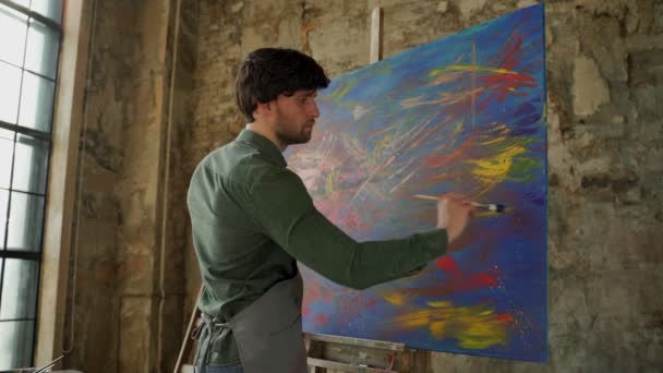Manlig konstnär målning bild i studio. Porträtt av en man som målar en bild med blå färg i en verkstad. — Stockvideo