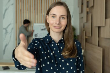 İş kadını kameraya elini uzatıyor, tokalaşıyor, misafirleri selamlıyor, kameraya bakıyor.