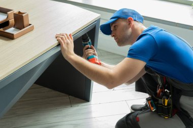 Erkek tamirci elinde elektrikli tornavida ile ahşap bir masayı tamir ediyor.