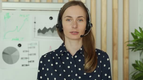 Młoda kobieta w słuchawkach, stojąca w biurze, pracująca jako operator call center lub support service, patrzy w kamerę — Wideo stockowe