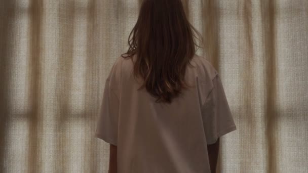 Taustakuva - nuori, hyväkuntoinen nainen valkoisessa t-paidassa avaa makuuhuoneen ikkunaverhon. — kuvapankkivideo