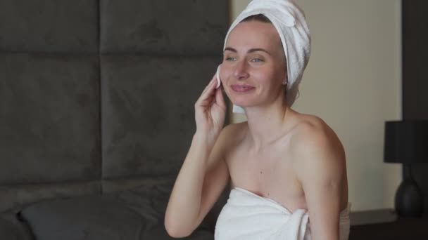 Молодая брюнетка с полотенцем на голове снимает макияж с лица, сидя на кровати. Концепция ухода за кожей и красоты. — стоковое видео