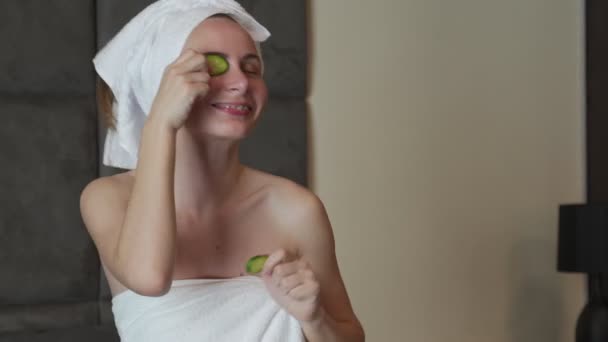 Femme avec une serviette sur la tête et des concombres devant ses yeux danse assis sur le lit. Prendre soin de la beauté à la maison — Video