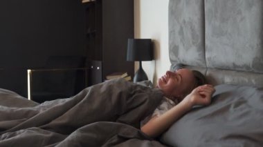 Beyaz tişörtlü bir kadın mutlu uyanıyor, esniyor, rahat bir yatakta oturuyor, modern bir otel odasında.
