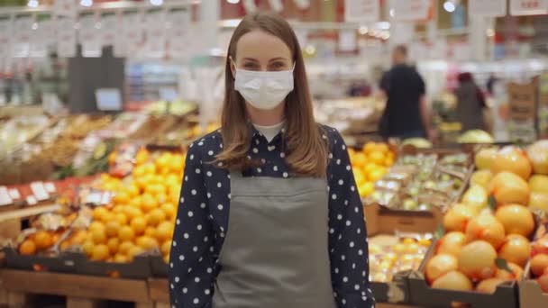 Портрет работницы в фартуке и защитной маске на фоне овощей и фруктов, молодая женщина скрещивает руки в продуктовом магазине и смотрит в камеру — стоковое видео