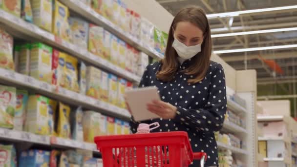 Kobieta shopper w masce ochronnej z wózkiem w przejściu sklepu z listy zakupów podczas zakupów żywności. Kobieta czyta w notatniku w nawie supermarketu. — Wideo stockowe