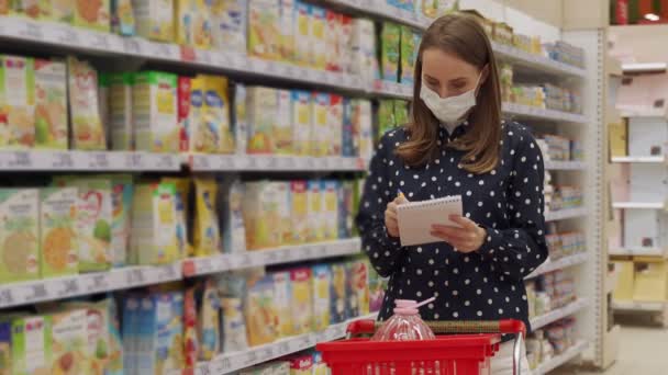 Kobieta shopper w masce ochronnej z wózkiem w przejściu sklepu z listy zakupów podczas zakupów żywności. Kobieta czyta w notatniku w nawie supermarketu. — Wideo stockowe