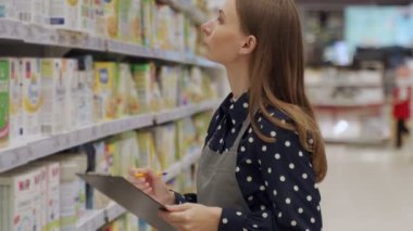 Süpermarkette çalışan önlüklü bir kadın. Bir kadın çalışan, marketin vitrinindeki ürünlerin envanterine bir tablet yazar..