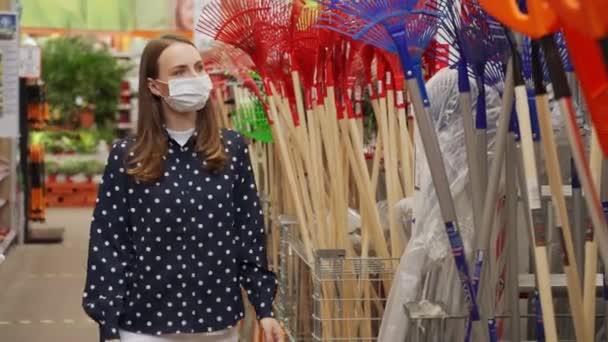 Mujer joven con máscara médica eligiendo un rastrillo en una tienda. Compra de artículos de jardín en una tienda departamental. — Vídeos de Stock