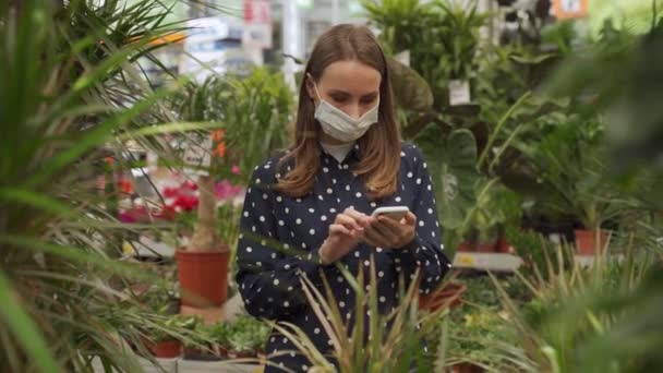Mujer con máscara médica comprando flores en un centro de jardinería usando un teléfono inteligente — Vídeos de Stock