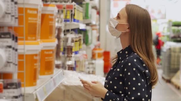 Woman wearing a protective mask picks up paint at a hardware store and uses a smartphone — Stock Video