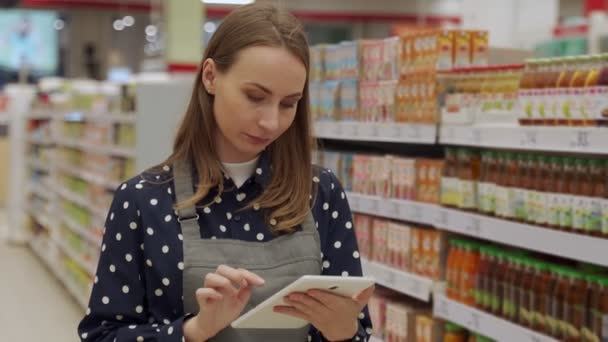 Uma mulher de avental a trabalhar num supermercado. Funcionária do sexo feminino usando um tablet digital para inventariar produtos em uma janela de supermercado. — Vídeo de Stock