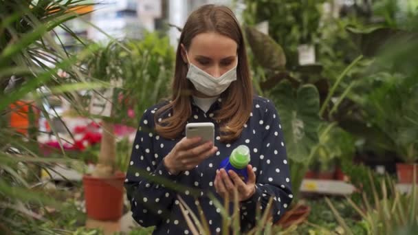 Tıbbi maskeli bir kadın, çiçek ve bitkiler için tarımsal kimyasalları akıllı telefon kullanarak seçiyor. — Stok video