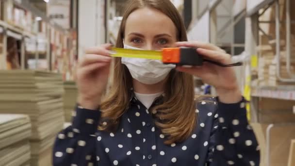 Mujer con una máscara protectora desenvuelve una cinta métrica de construcción en una tienda de construcción — Vídeo de stock
